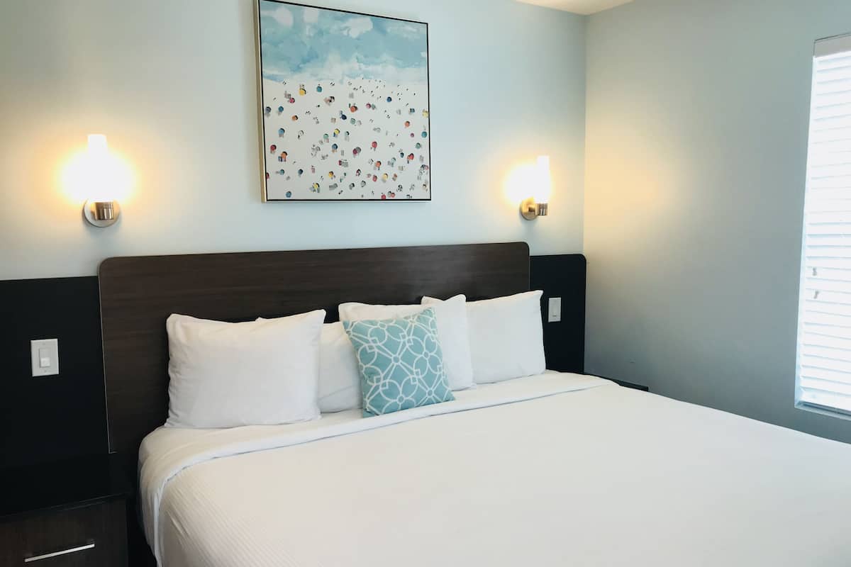 A hotel room with a large bed, two wall-mounted lights, a painting above the bed, and a window on the right.