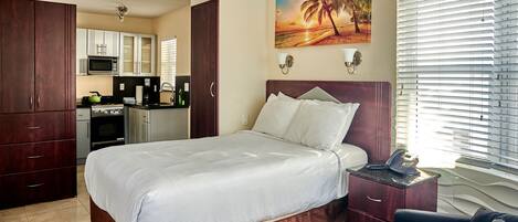 A hotel room with a bed, a desk with a phone, a chair, a painting of a beach scene, and a kitchenette in the background.