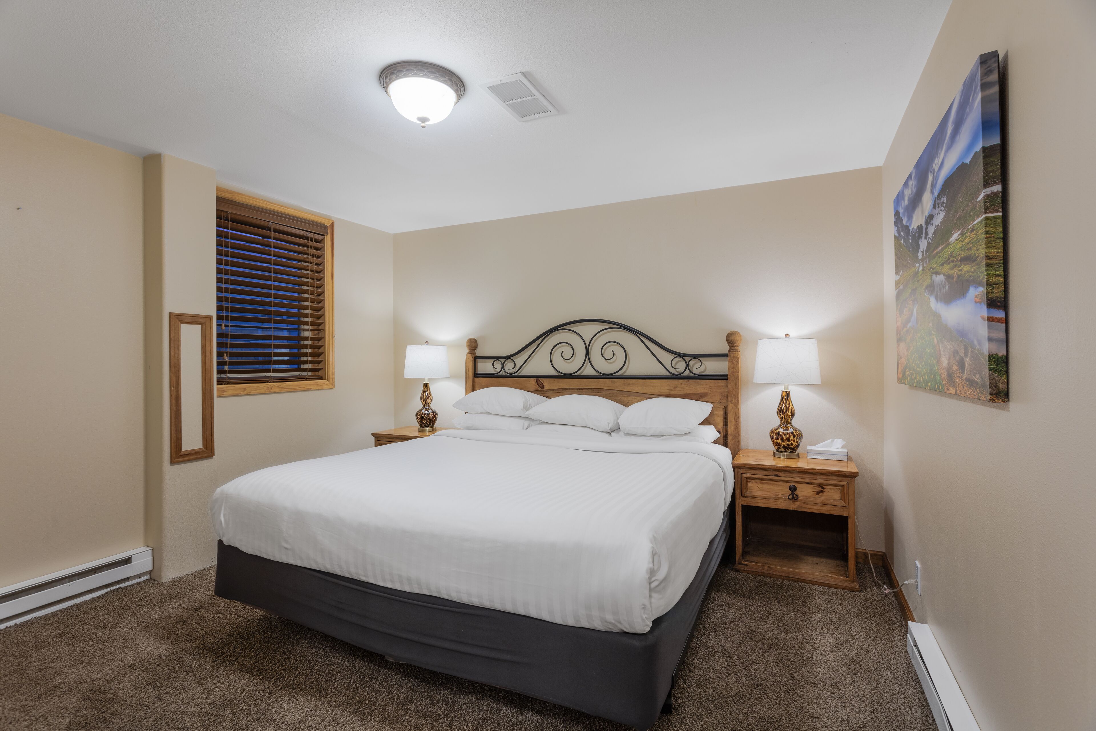 A bedroom with a large bed, two bedside tables with lamps, a window with blinds, and a painting on the wall.