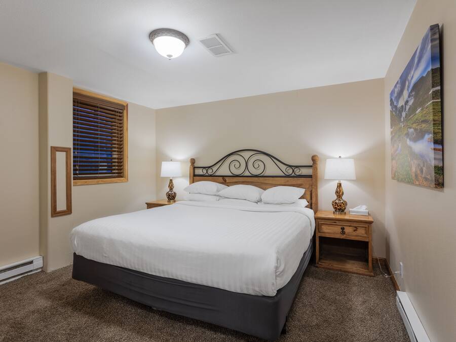 A bedroom with a large bed, two bedside tables with lamps, a window with blinds, and a painting on the wall.