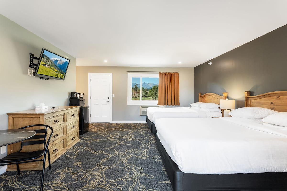 A hotel room with two beds, a desk, a chair, a television mounted on the wall, and a window with a view of mountains.