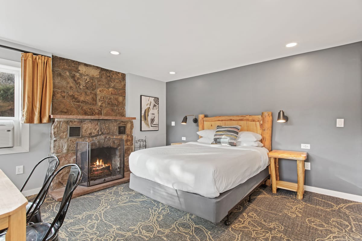 A modern bedroom with a stone fireplace, a bed with white linens, and a small wooden nightstand.