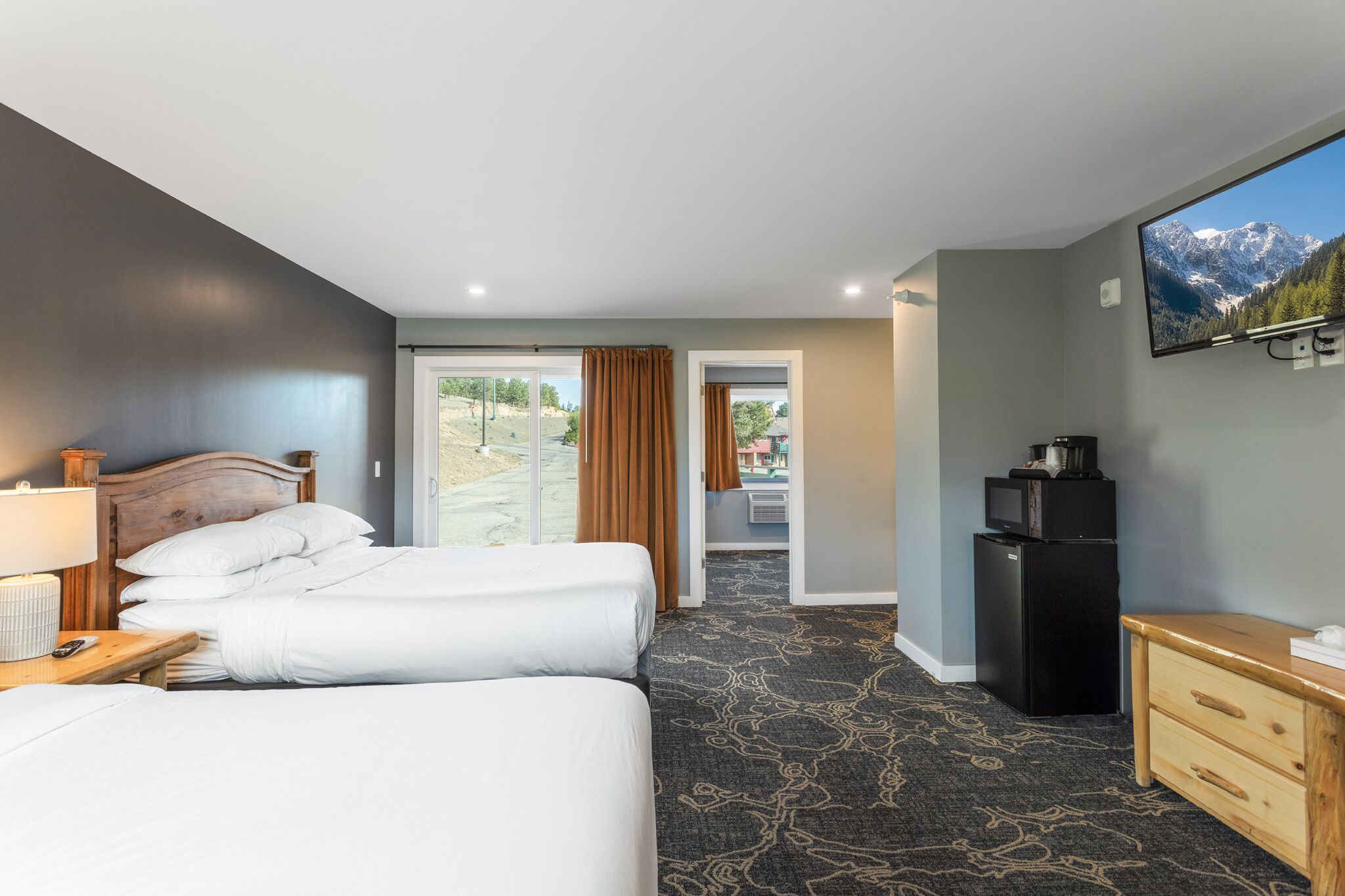 A hotel room with two beds, a TV, a refrigerator, and a view of mountains through the window.