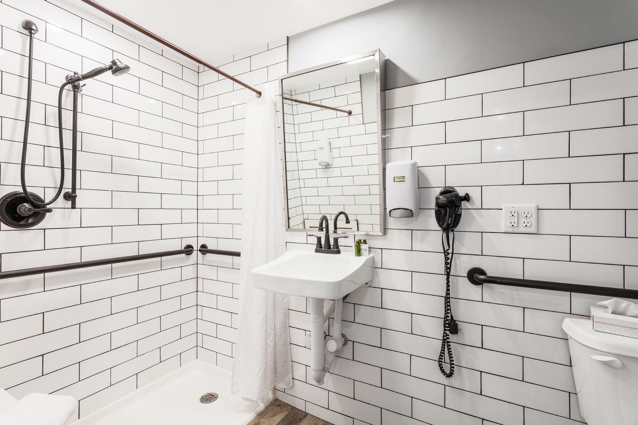 A modern bathroom with white subway tiles, a white sink, a handheld showerhead, a towel rack, a toilet, and a telephone.