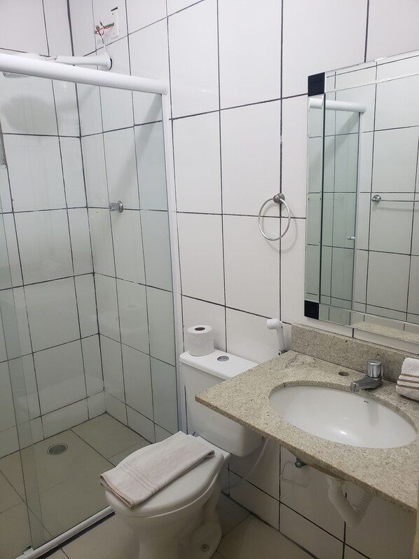 A bathroom with white tiled walls, a toilet, a sink with a granite countertop, and a shower area. - Pousada Beira Mar (Santos)