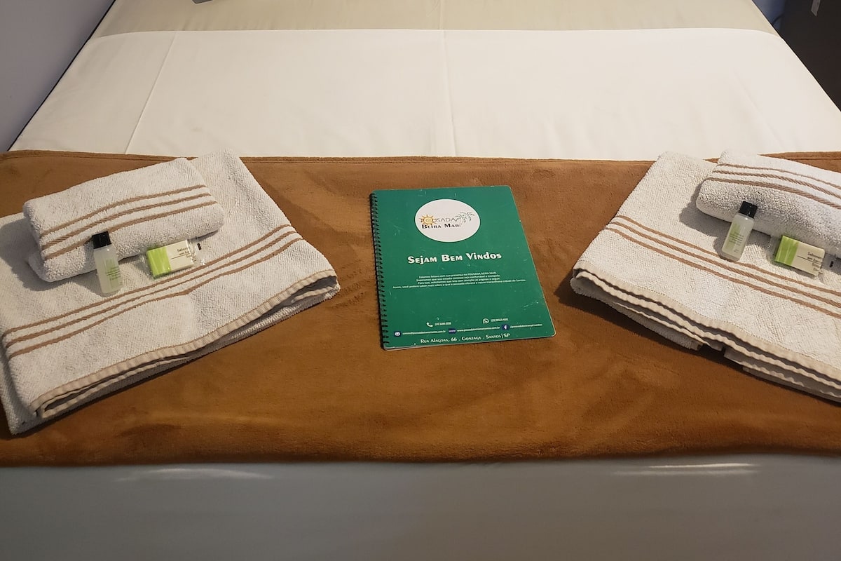 Ein ordentlich bezogenes Bett mit zwei Kissen, einem grünen Buch mit der Aufschrift 'Sejam Bem Vindos' und zwei Handtüchern mit Toilettenartikeln auf einer braunen Bettdecke.