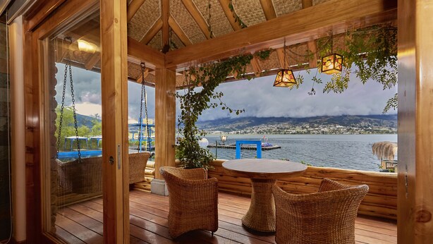 A wooden deck with wicker furniture, a view of a lake, and a cityscape in the background. - Hostería Cabañas del Lago (San Pablo del Lago)