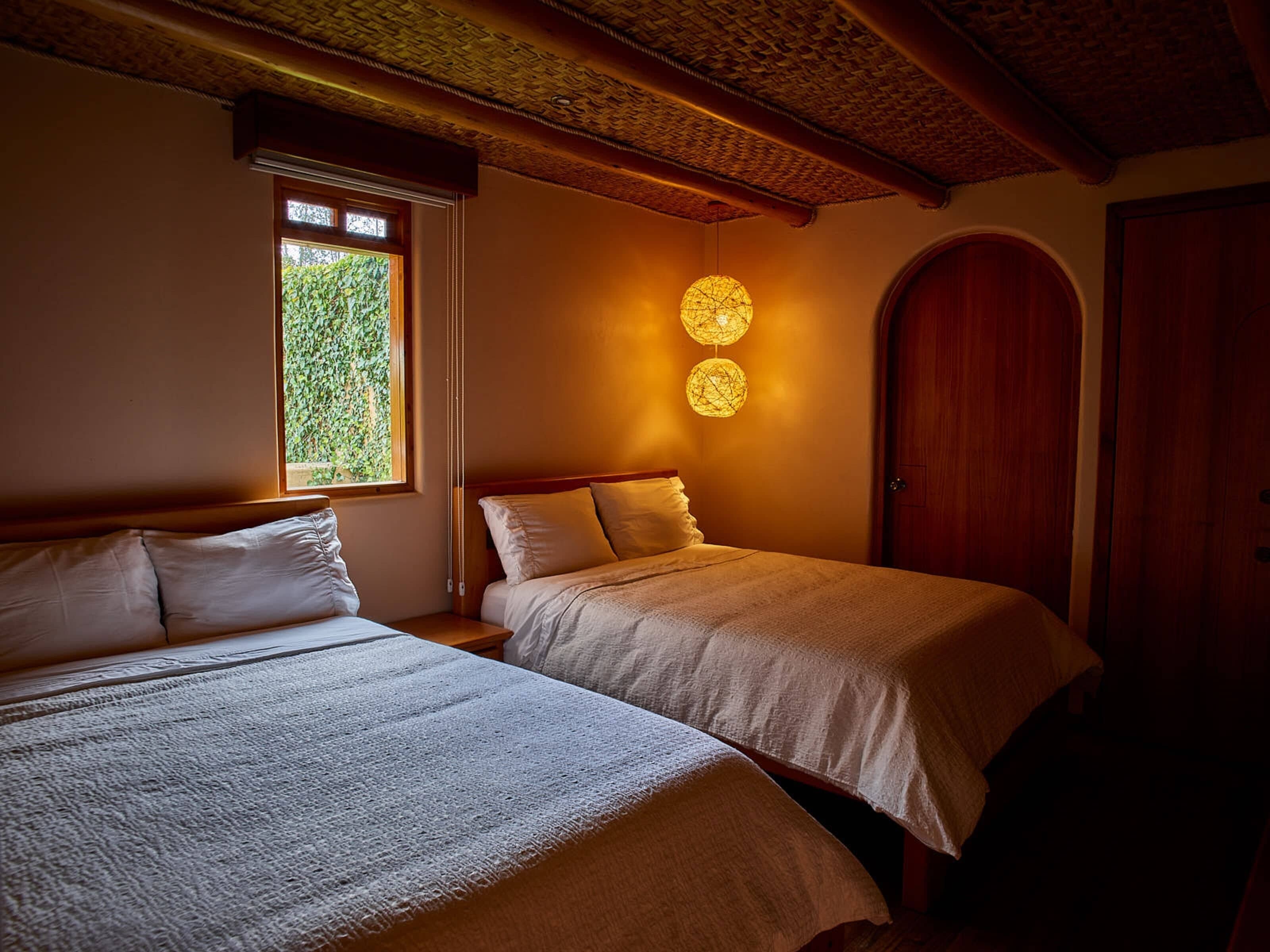 Quarto com duas camas, janela com vista para vegetação, luminária esférica fixada na parede e porta de madeira.