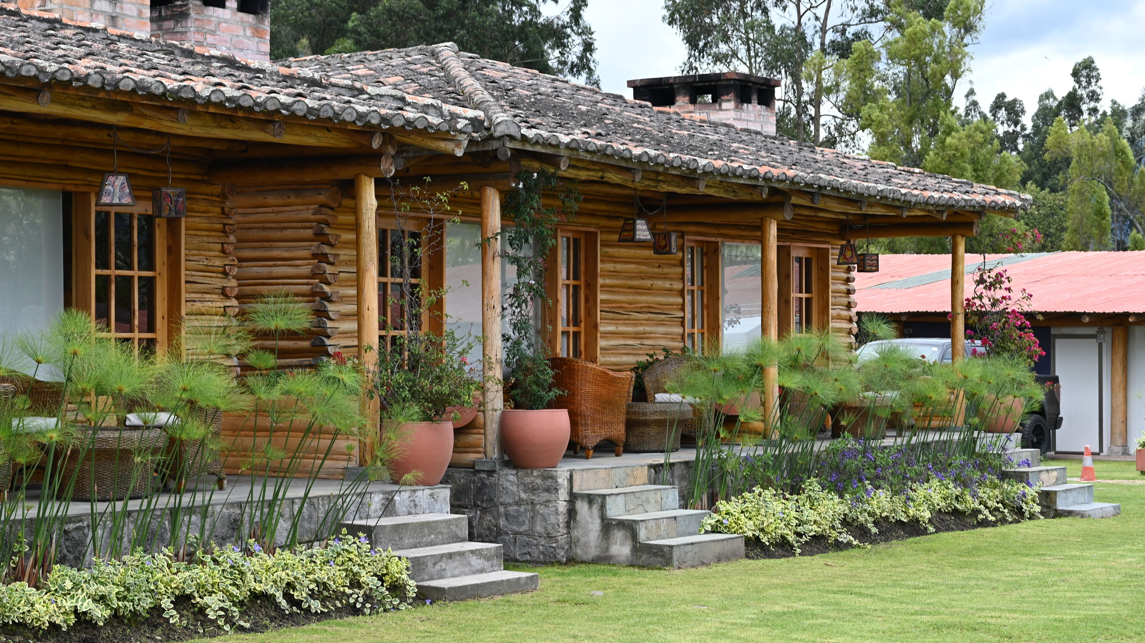 A rustic log cabin with a tiled roof, large potted plants, and a stone staircase leading to the entrance.