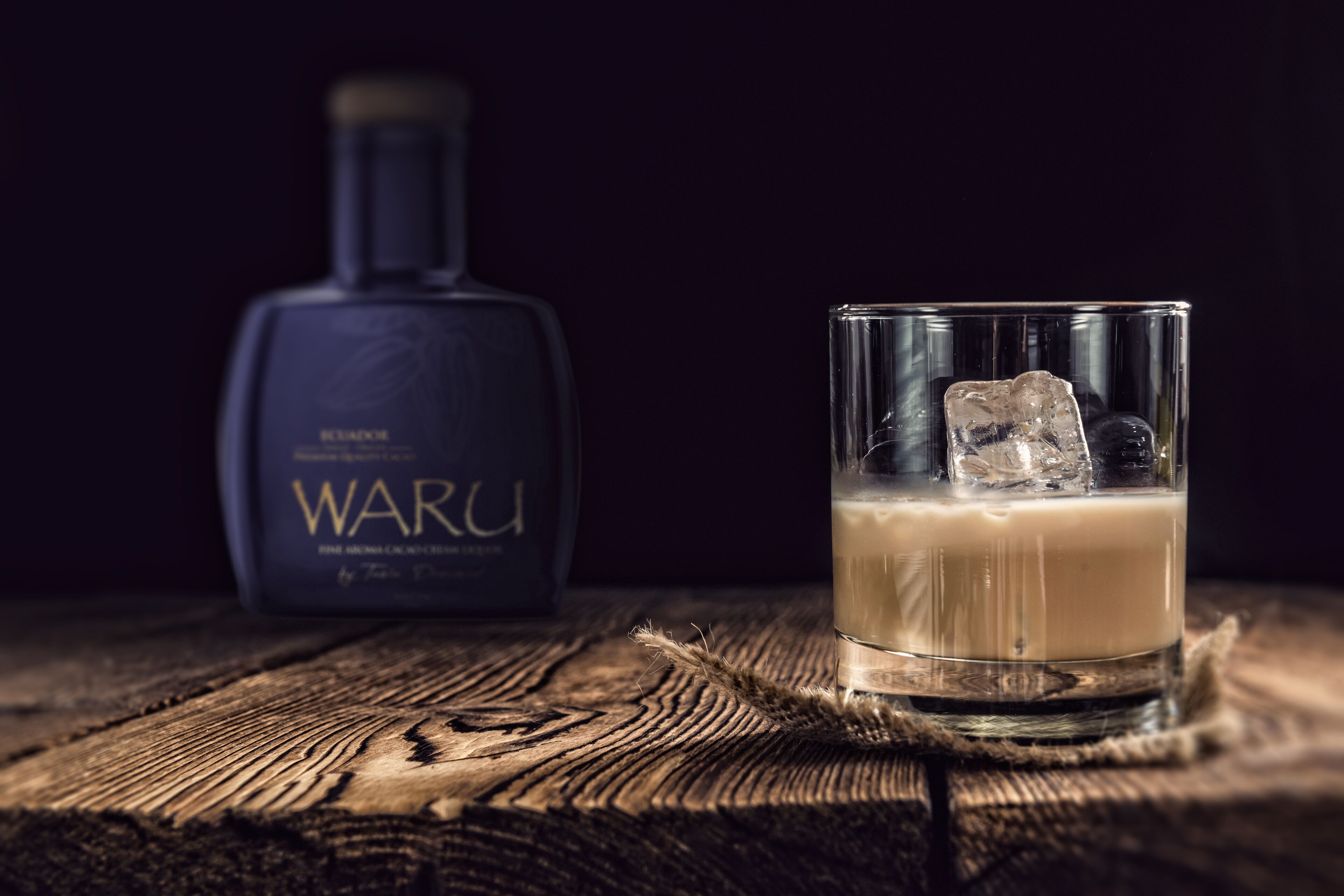 A glass of a beverage with ice cubes on a wooden surface, a bottle labeled "WARU" in the background.
