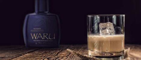 A glass of a beverage with ice cubes on a wooden surface, a bottle labeled "WARU" in the background.