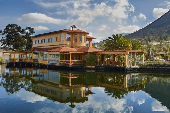 A large, multi-story house with a prominent tower, situated by a calm body of water, reflecting the structure. - Hostería Cabañas del Lago (San Pablo del Lago)