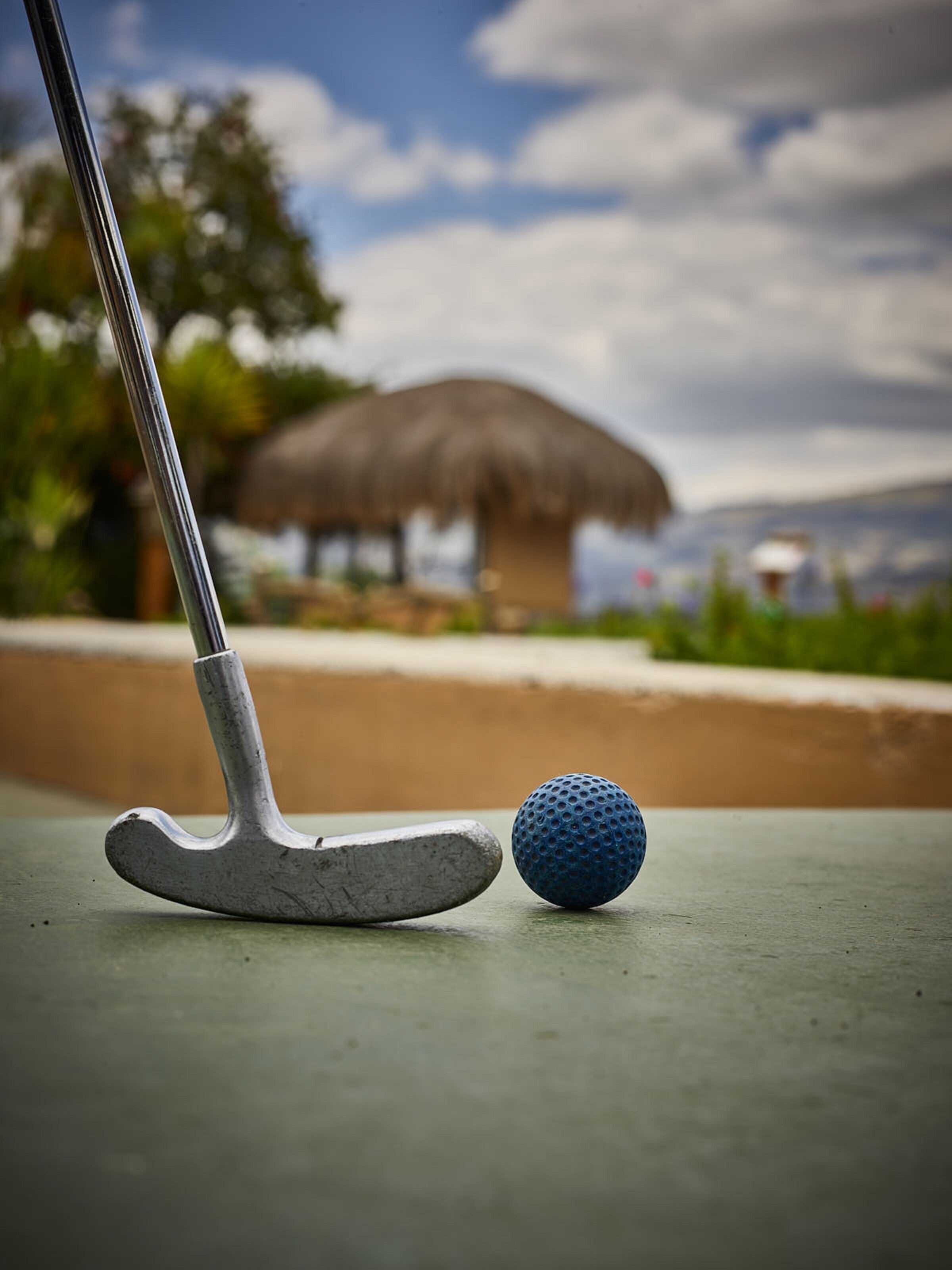 A putter and a golf ball on a green surface with a thatched-roof structure in the background.