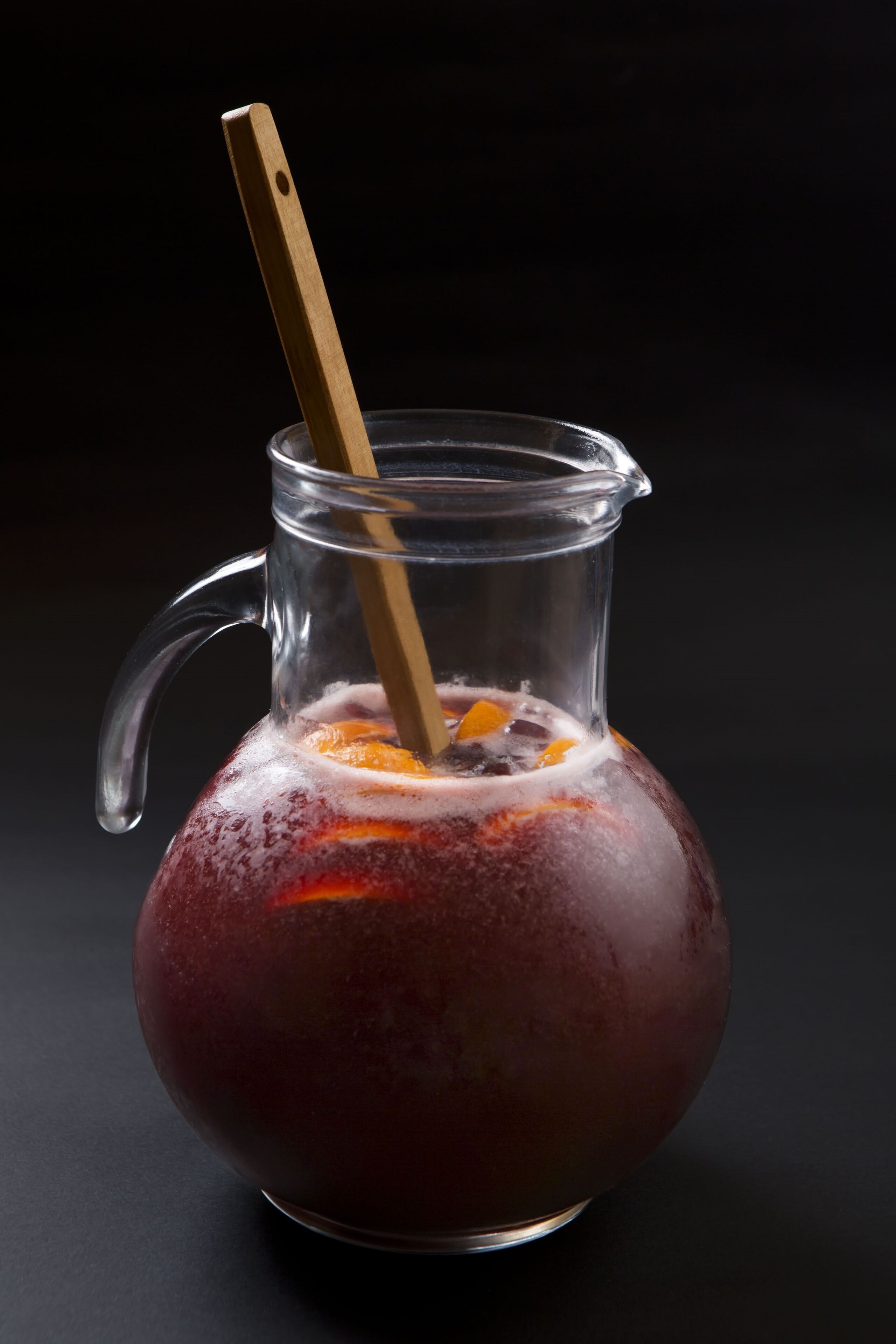 A clear glass pitcher with a wooden handle filled with a red beverage and ice cubes, placed on a dark surface.