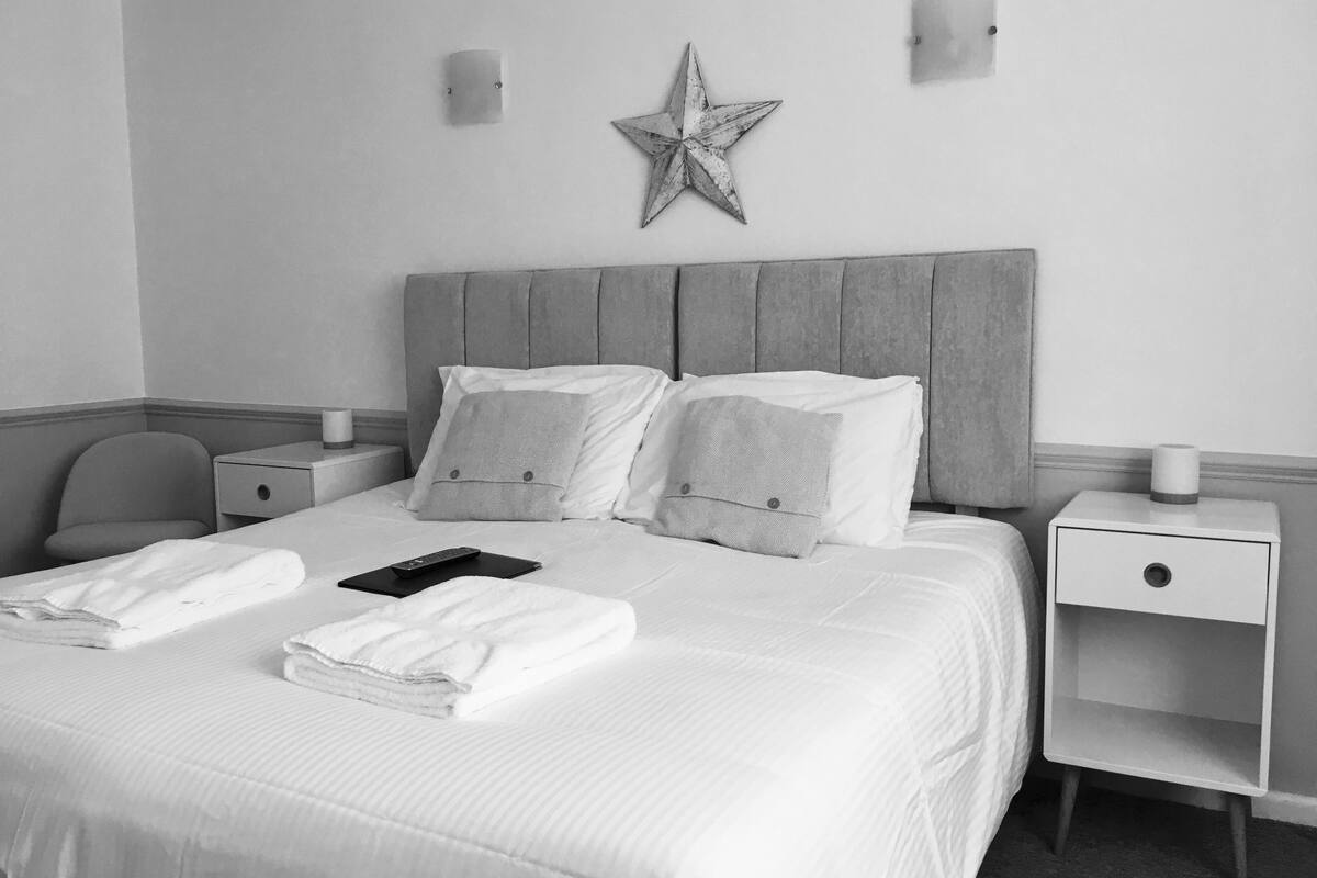 A neatly made bed with pillows, a remote control, and folded towels. There are two bedside tables, one with a lamp and the other with a cup. A star decoration hangs on the wall above the bed.