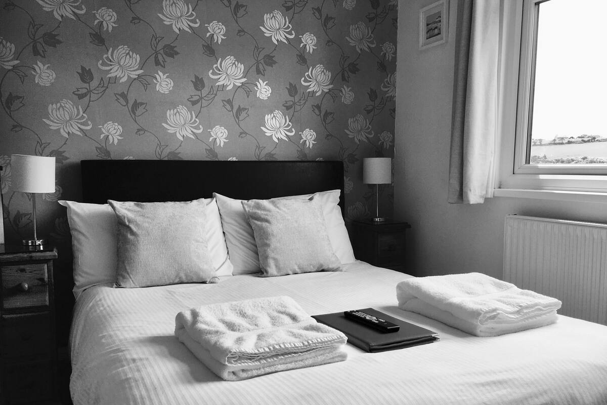 A neatly made bed with pillows and folded towels, a remote control, and a lamp on a bedside table.