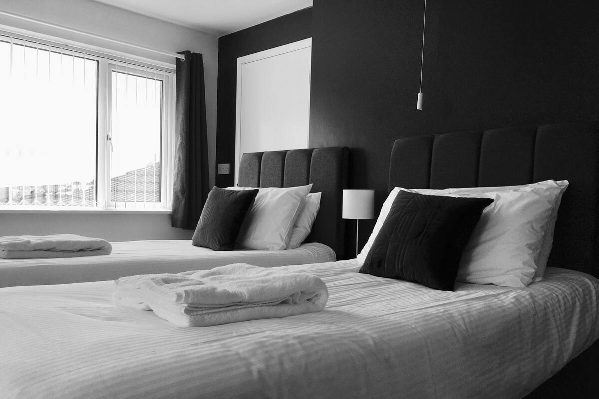 A hotel room with two beds, a large window with blinds, a bedside lamp, and a view of the cityscape.