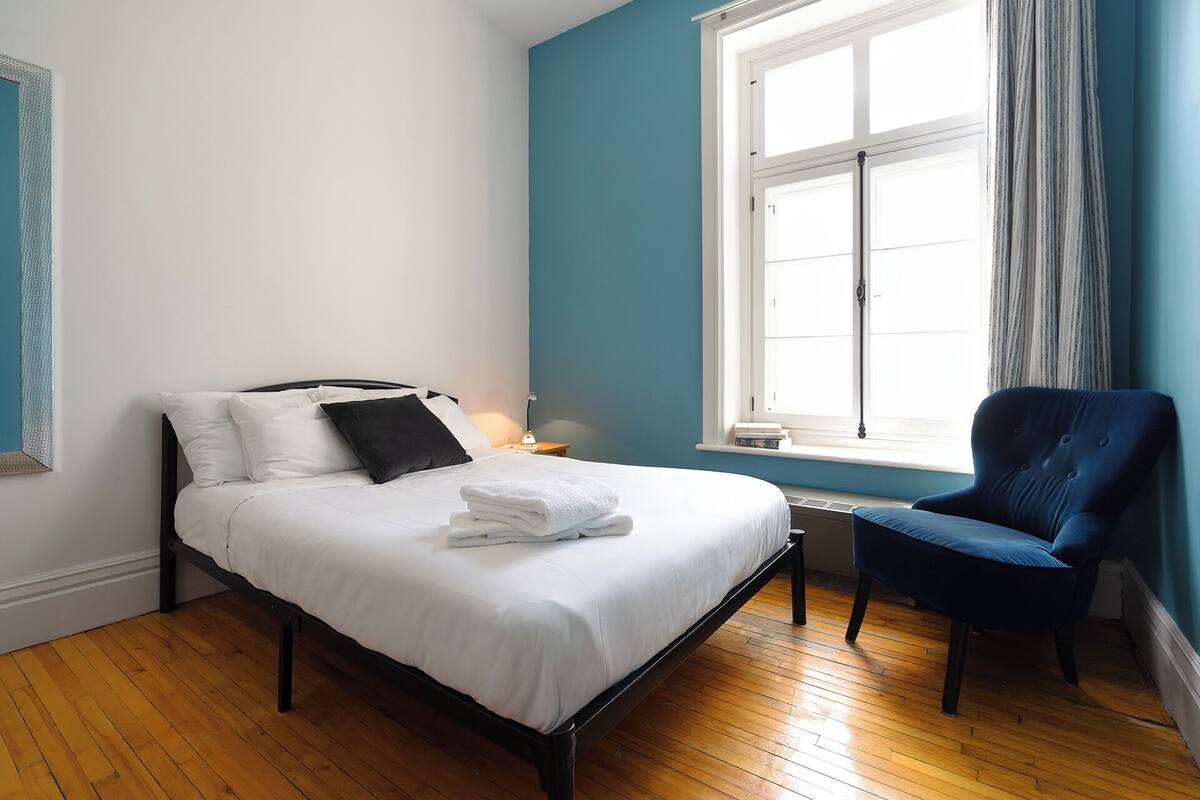 Un dormitorio con cama, una silla, una ventana y una mesita de noche.
