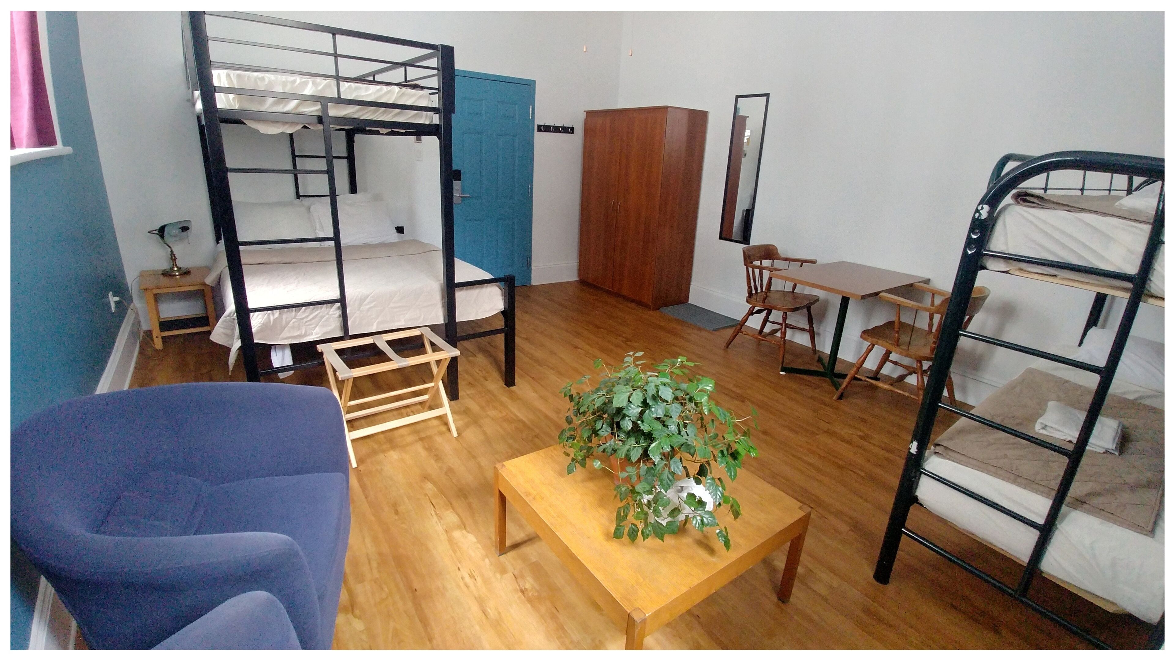 A dormitory room with bunk beds, a wooden table, a chair, a desk, a mirror, and a wardrobe.