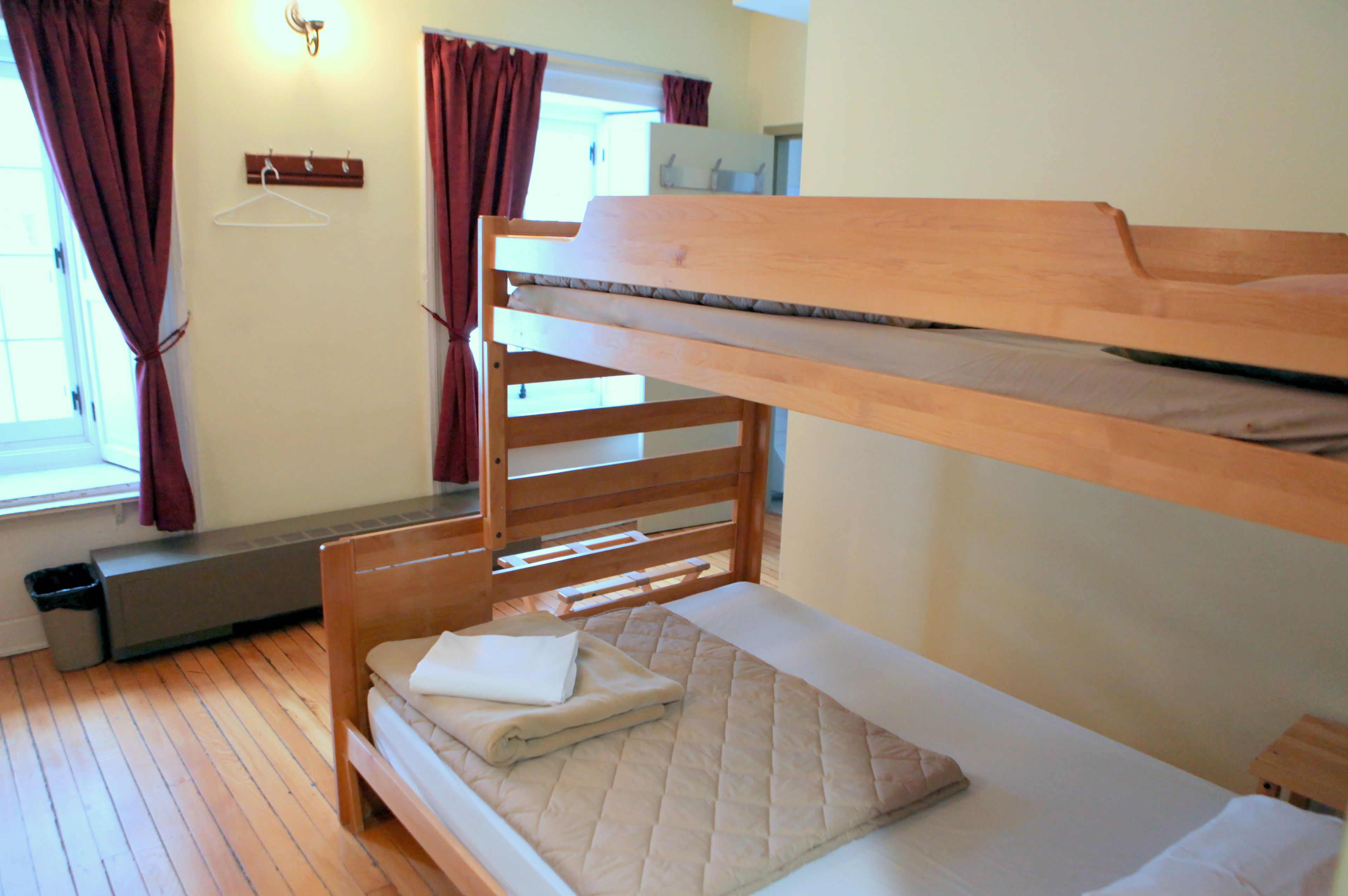 A bunk bed room with a wooden bunk bed, a lower bed with a quilted mattress, a bench, a radiator, and a window with curtains.