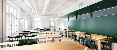A classroom with tables and chairs, a large green chalkboard, and windows allowing natural light.