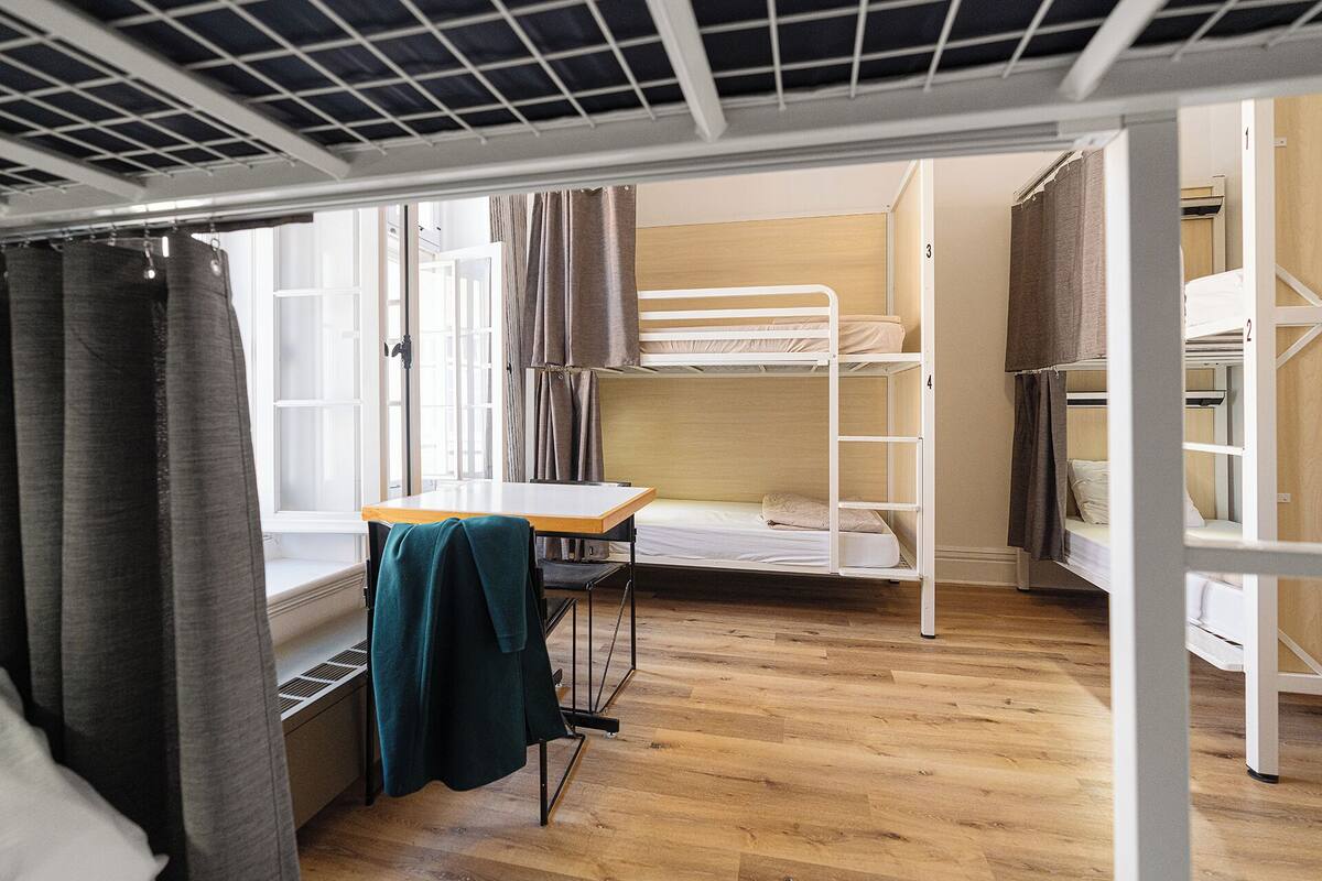 Una habitación pequeña con literas, una mesita y una ventana con cortinas.