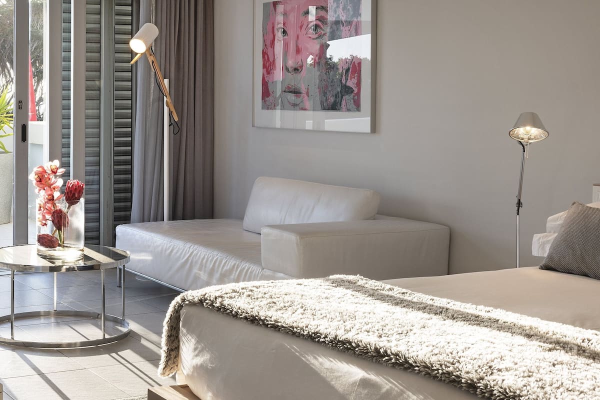 A modern hotel room with a sofa, a bed, a round table with a vase of flowers, and a painting on the wall.