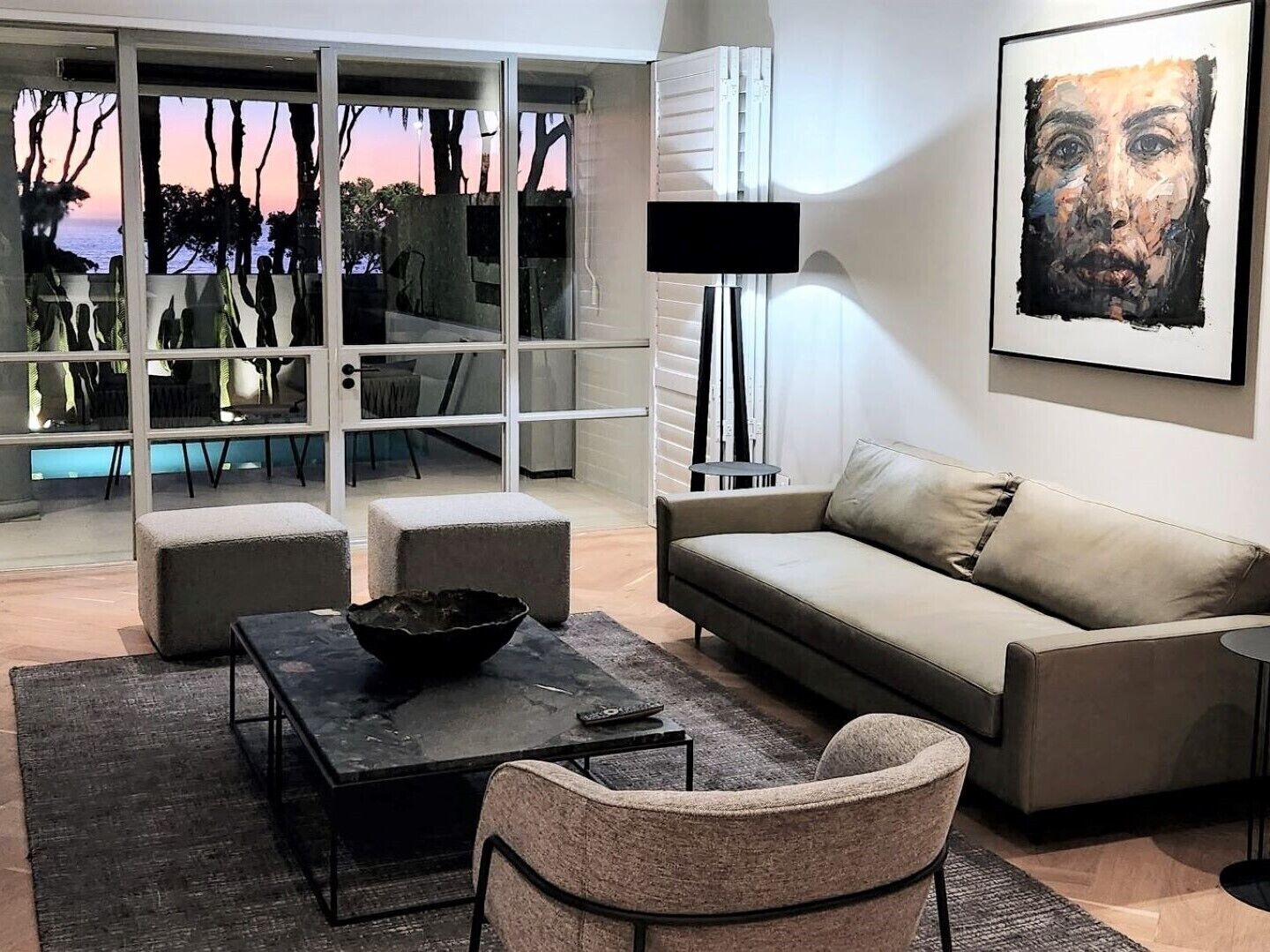 Un salon moderne avec un canapé, une table basse et un fauteuil. Une grande fenêtre offre une vue sur les arbres et un coucher de soleil.