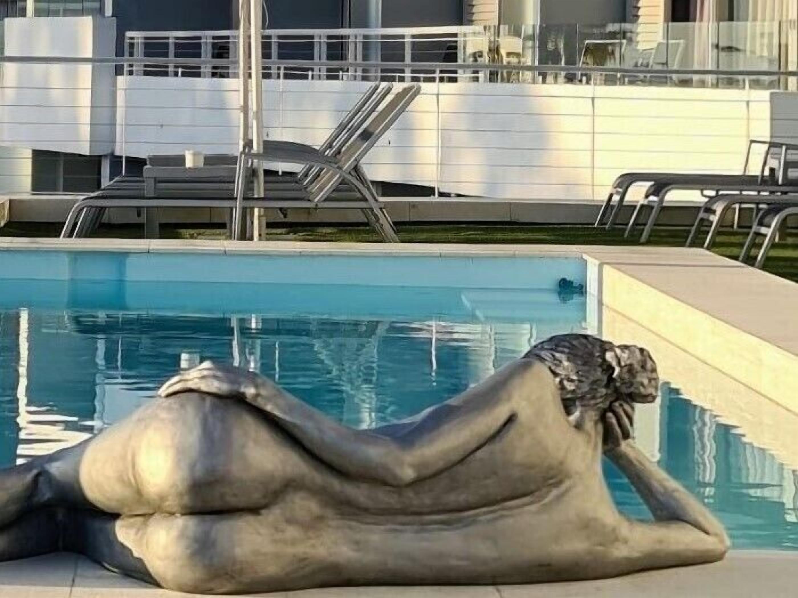 A sculpture of a reclining woman by a pool with a modern building in the background.