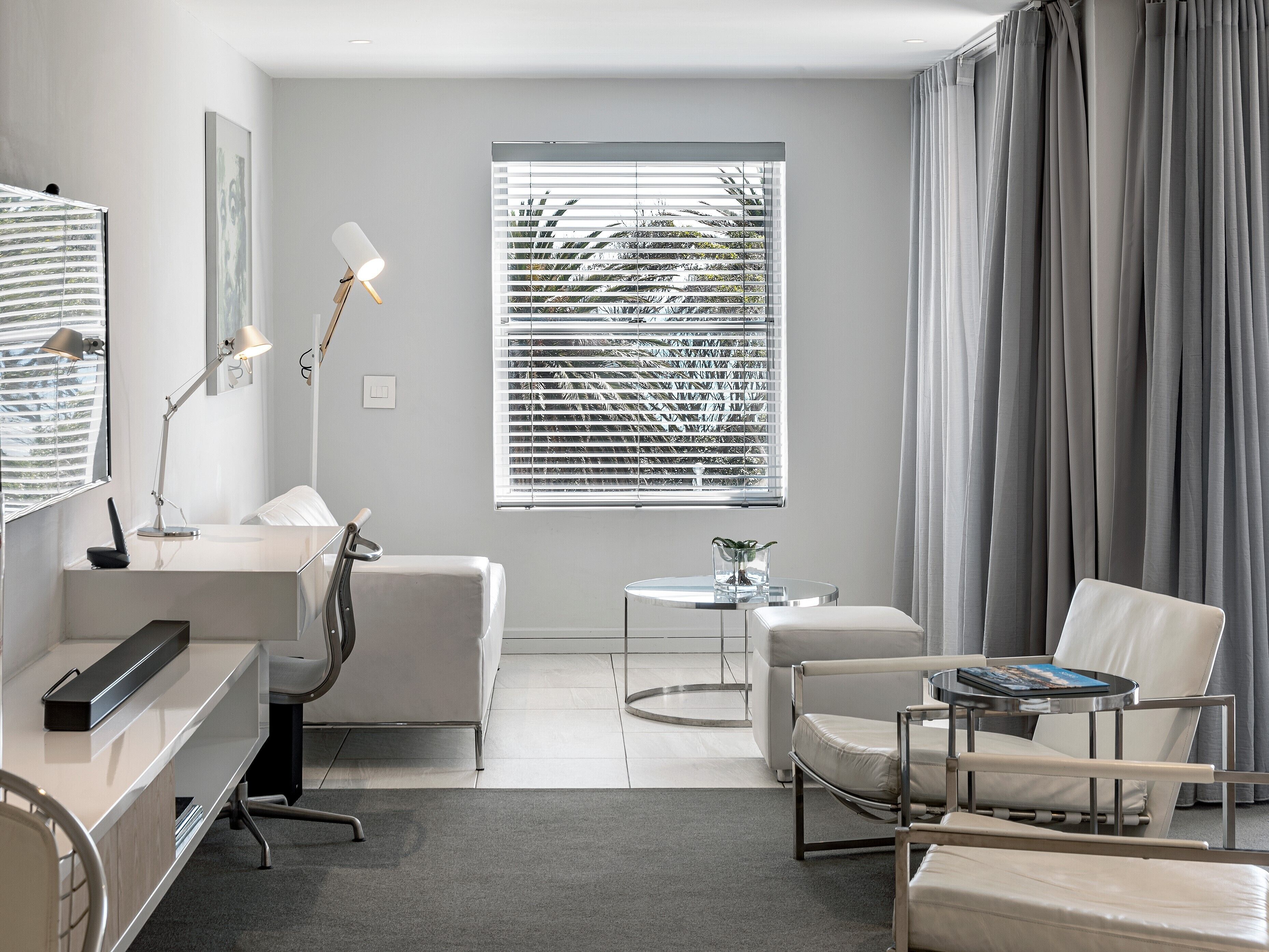 A modern hotel room with a desk, two chairs, a small table, a mirror, and a window with blinds.
