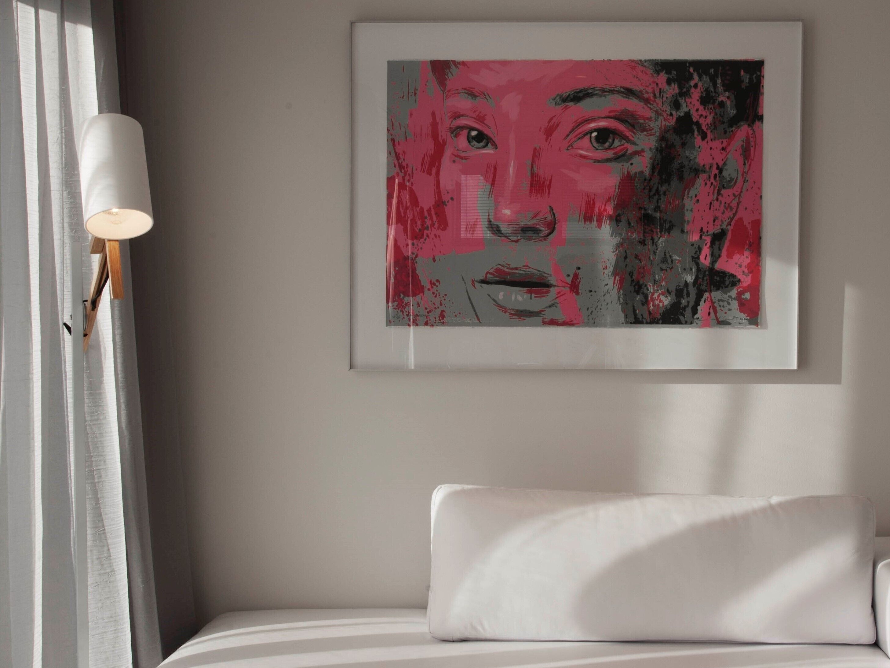 A framed artwork on a wall above a bed with a white pillow and a white lamp on a bedside table.