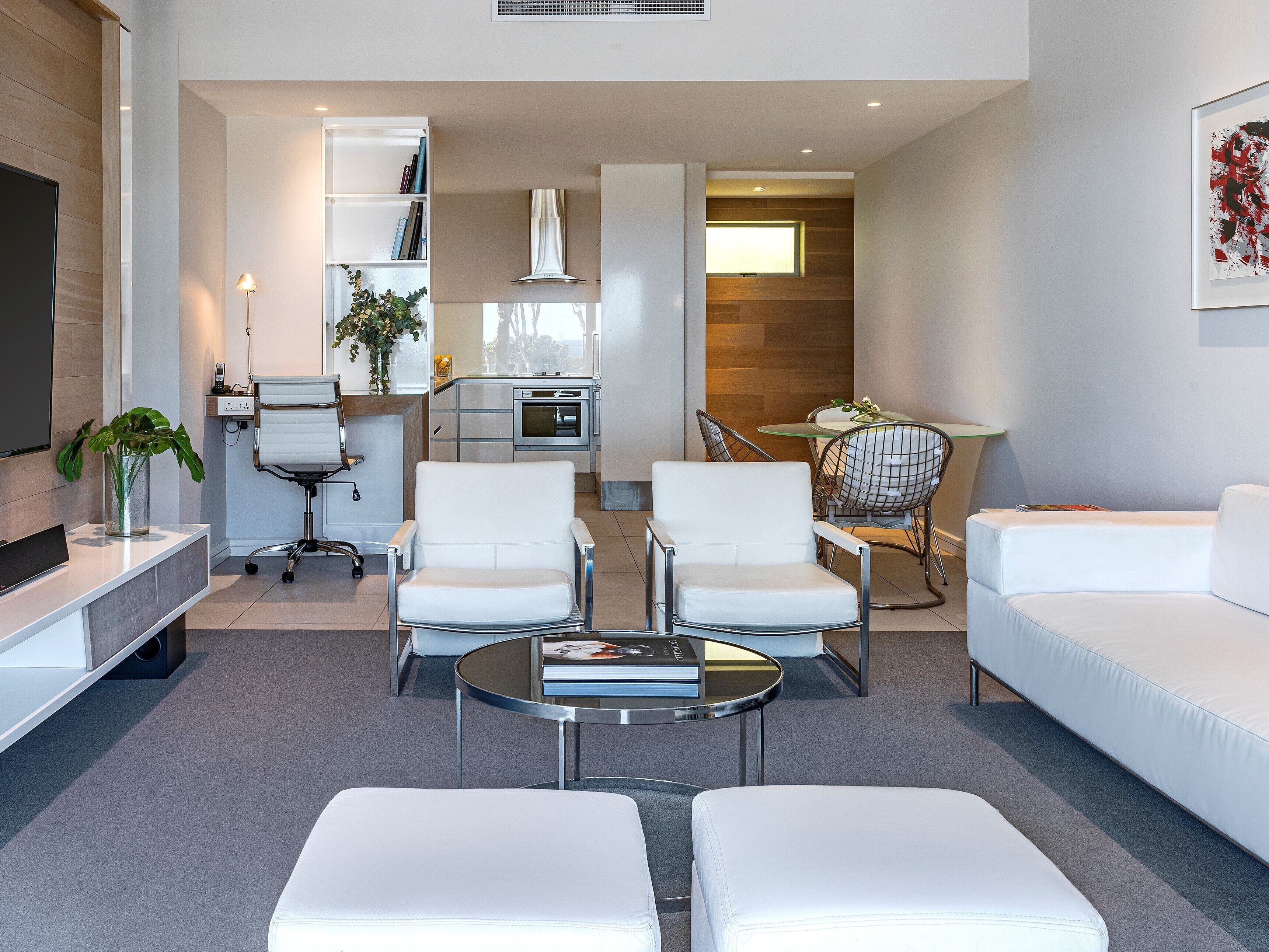 A modern living room with a sofa, two armchairs, a coffee table, and a TV. There is a kitchen area with a stove and a dining table with chairs.