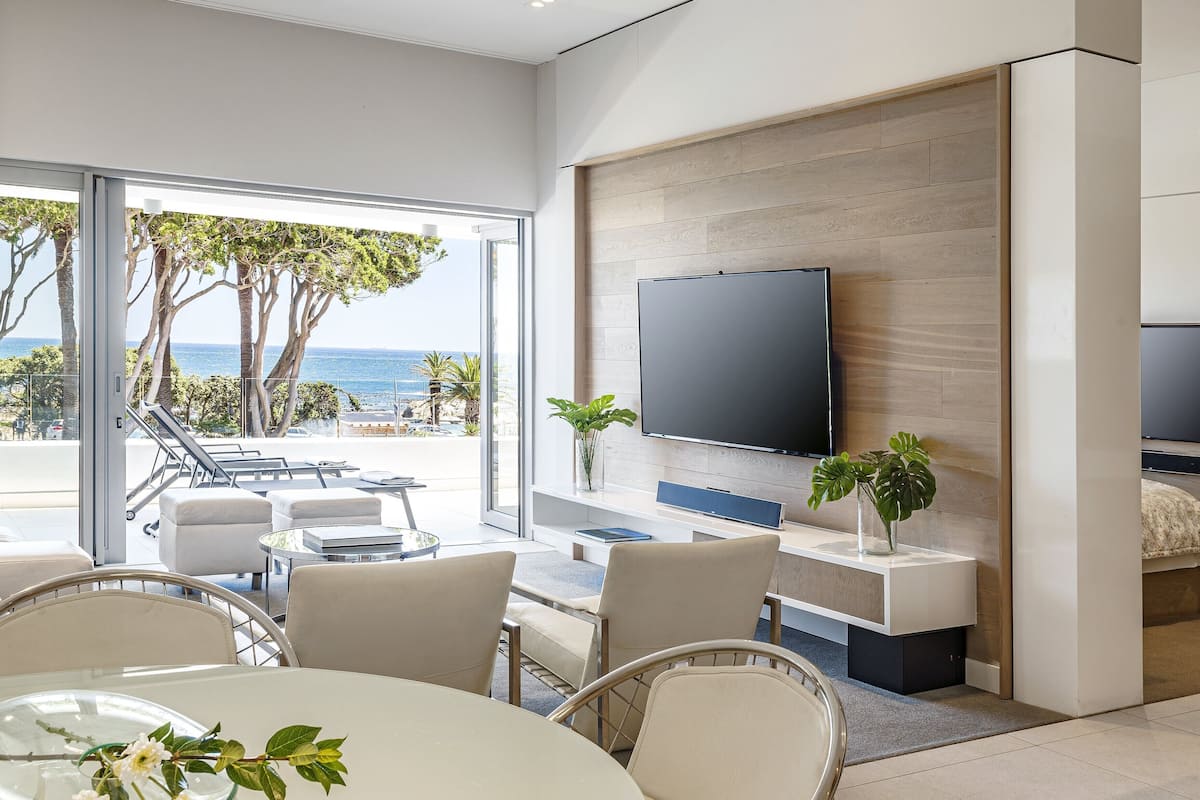A modern living room with a large TV, comfortable seating, a dining area, and a view of the ocean.