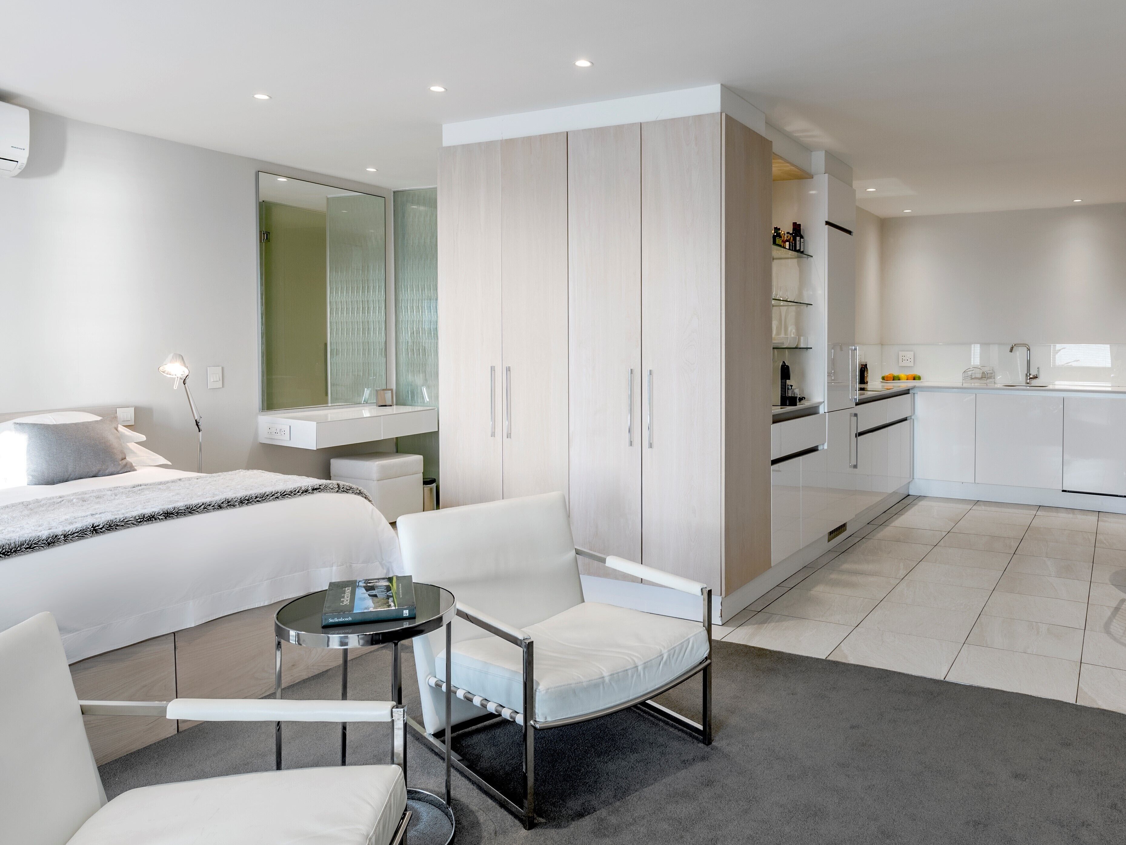 A modern hotel room with a bed, a sitting area with a chair and table, a wardrobe, a bathroom with a mirror, and a kitchenette.