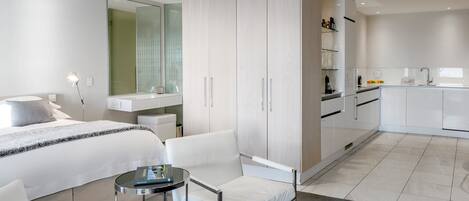 A modern hotel room with a bed, a sitting area with a chair and table, a wardrobe, a bathroom with a mirror, and a kitchenette.