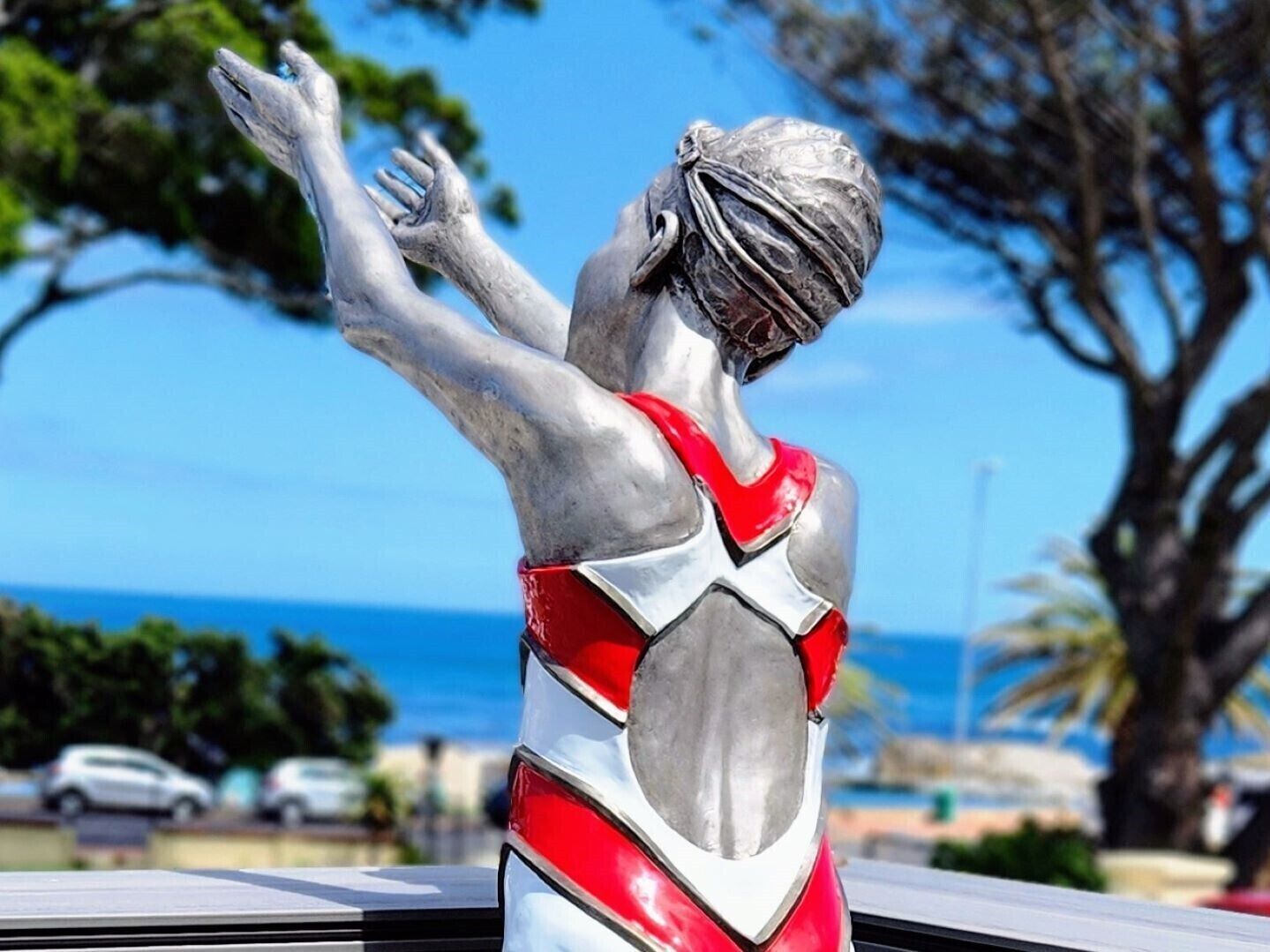 Une statue représentant une figure vêtue d’une tenue rouge et blanche, dans une pose dynamique, se dressant sur un fond composé d’arbres et d’un ciel dégagé.