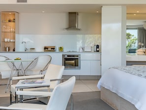 A modern hotel room with a kitchenette, a dining area with a glass table and chairs, and a large bed with a view of the outdoors.