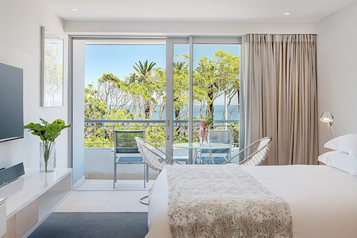 A modern hotel room with a large bed, a glass table with chairs, a TV, and a balcony with a view of trees and the sea.
