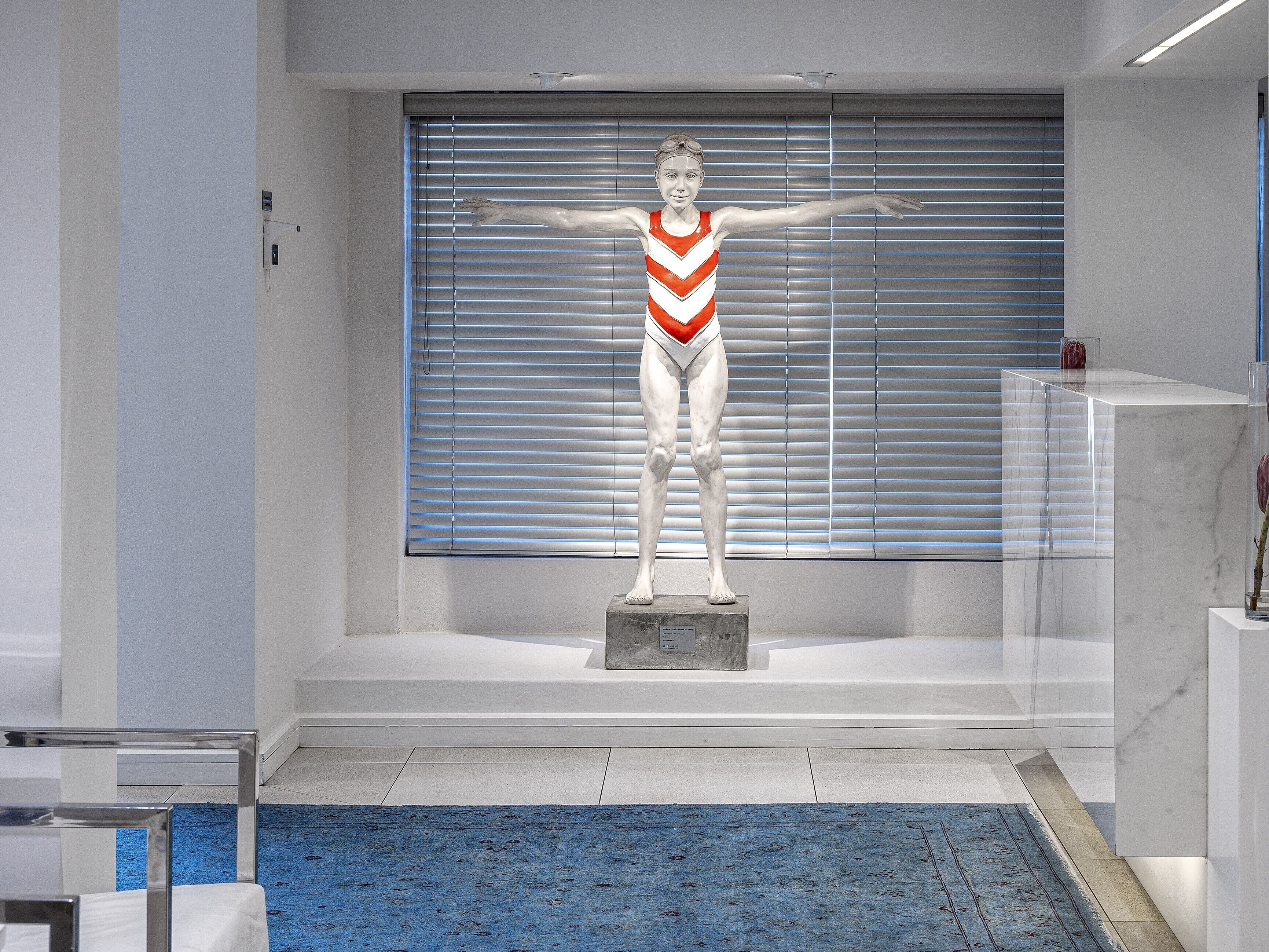 A statue of a swimmer in a white and red swimsuit on a pedestal in a modern lobby with a blue carpet and a large window with blinds.