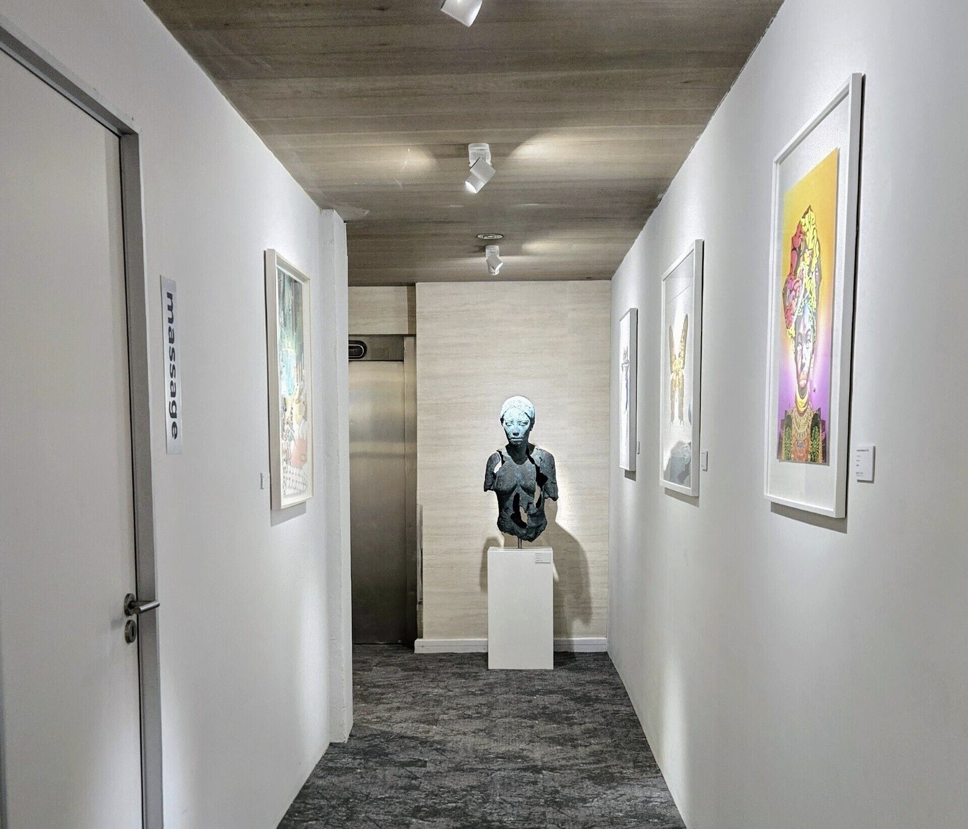 A hallway with a bust sculpture on a pedestal, framed artwork on the walls, and a carpeted floor.