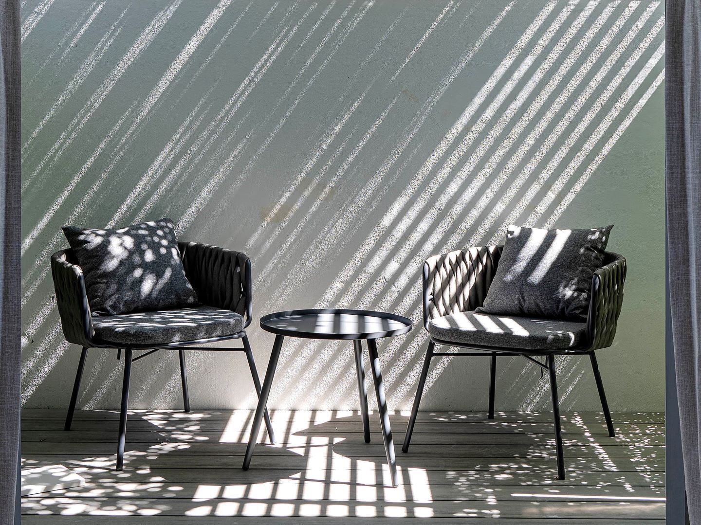 Two armchairs with patterned cushions and a small round table in a sunlit room with a textured wall.