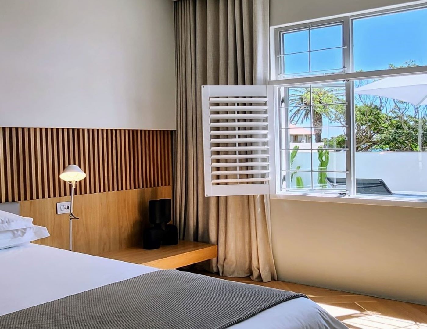 A hotel room with a bed, a bedside table, a lamp, a window with curtains, and a view of the outside.