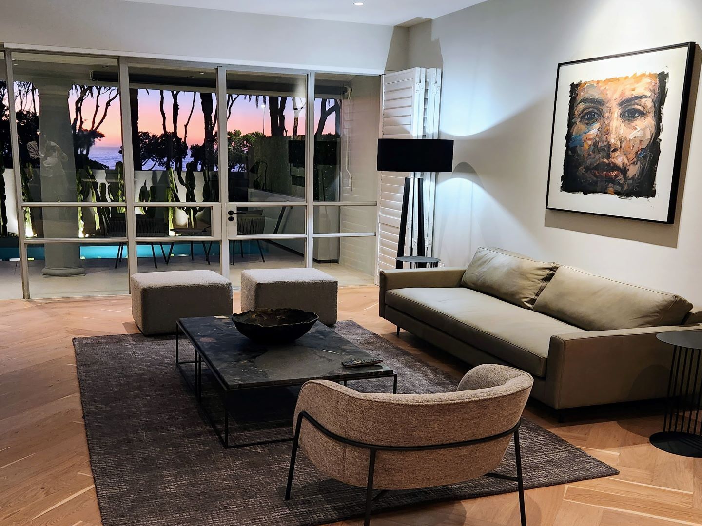 A modern living room with a sofa, coffee table, and armchair, large windows with a view of trees and sky, and a painting on the wall.