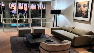 A modern living room with a sofa, coffee table, and armchair, large windows with a view of trees and sky, and a painting on the wall.