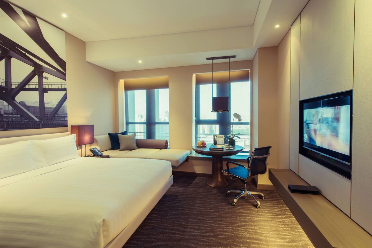 A hotel room with a large bed, a desk with a chair, a TV, and large windows.