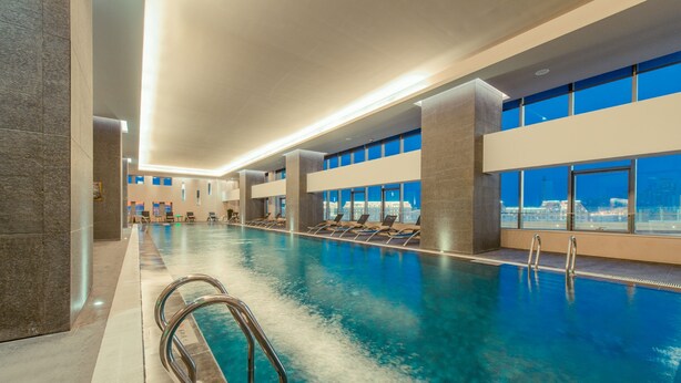 Una amplia piscina cubierta con agua cristalina, varias zonas de descanso con sillones y grandes ventanales con vistas a la ciudad.