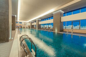 Une grande piscine intérieure à l’eau cristalline, plusieurs espaces de détente avec des fauteuils et de grandes fenêtres offrant une vue sur la ville.