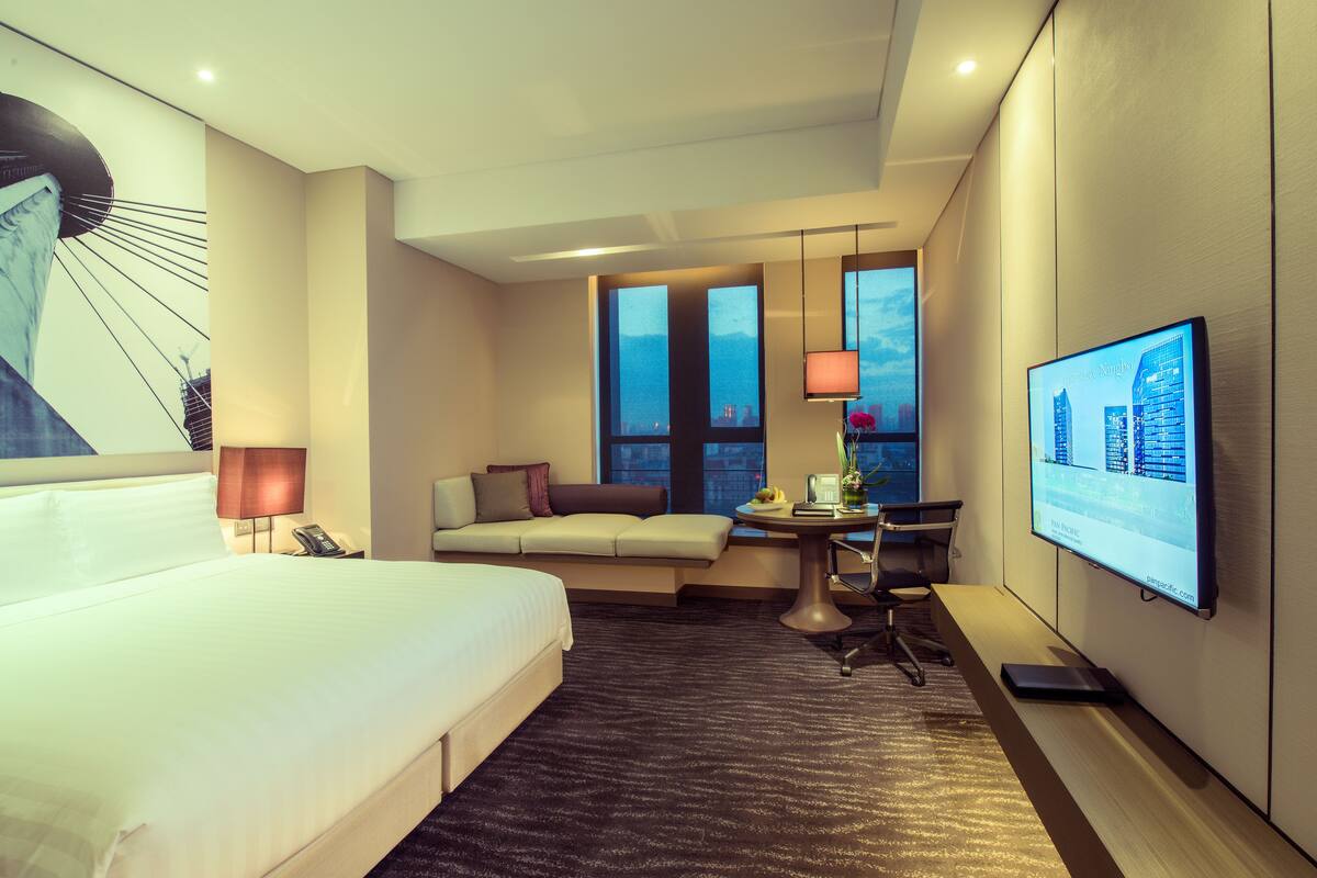 A hotel room with a large bed, a sofa, a desk with a computer, and a TV on the wall.