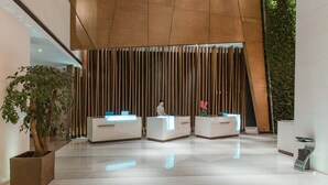 Un área de recepción moderna con tres mostradores blancos, una planta en maceta grande y un diseño de paneles de madera verticales.
