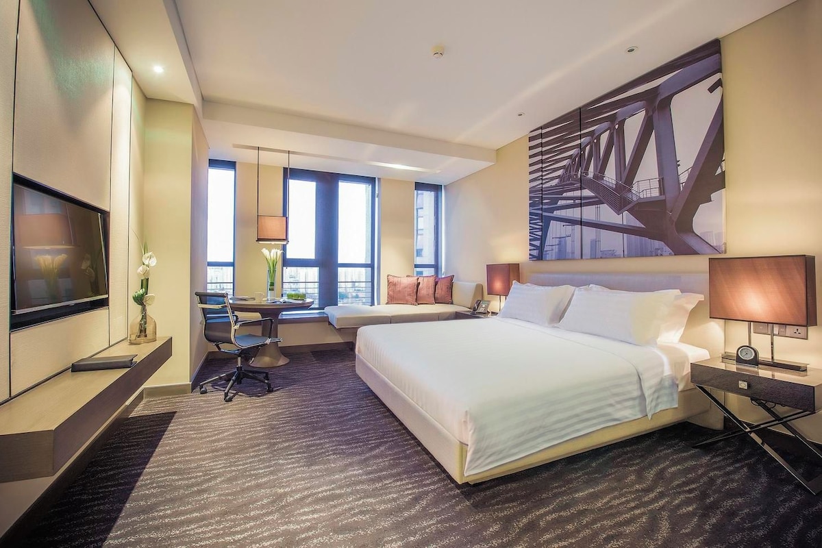 A hotel room with a large bed, a desk with a chair, a television, and a large window.